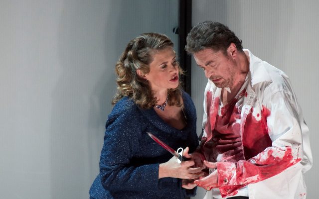 Lady in a blue suit stands next to a man in a blood-stained shirt holding a dagger