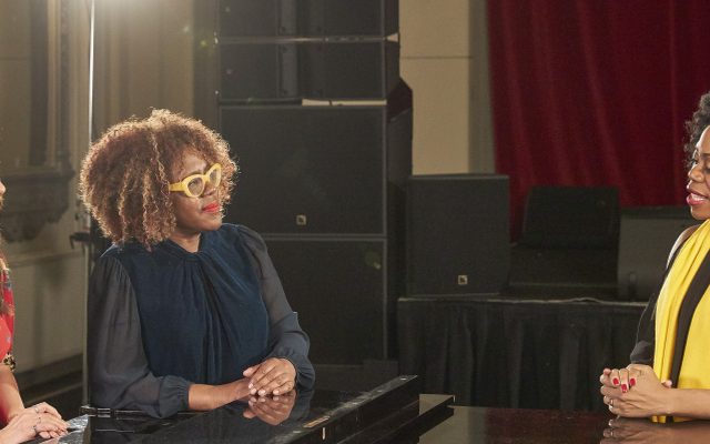 Three women stood at a piano talking to each other.