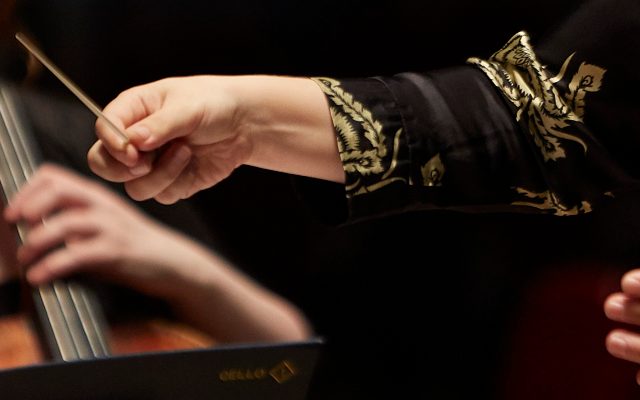 A woman's hands in ornate sleeves conducting an orchestra using a baton, with a cellist in the background