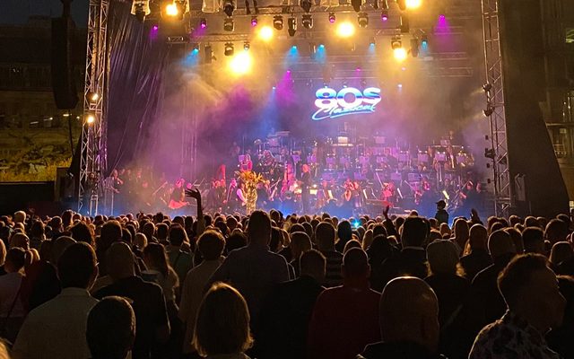 An orchestra and singers on a distant, bright;y lit stage, seen from the midst of a large standing audience