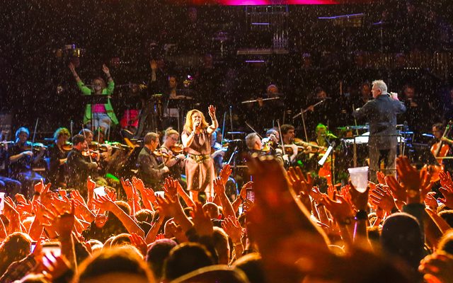 With hands in the air a crowd applauds a woman performing in front of an orchestra on a large open-air stage