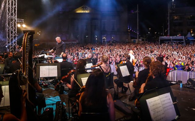A large outdoor crowd seen from the back of an orchestra onstage, with a conductor leading them