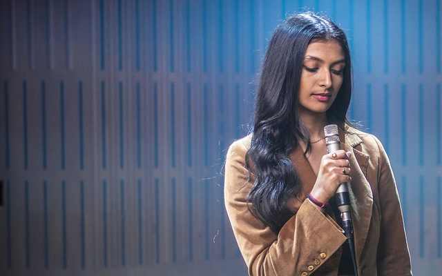 A young woman holding a microphone and looking down, lit from above by a blue haze, with wooden panelling behind her