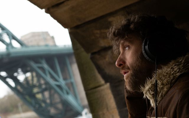 A bearded man wearing headphones and an Afghan jacket, seen in profile under a stone bridge, with a steel bridge visible in the background
