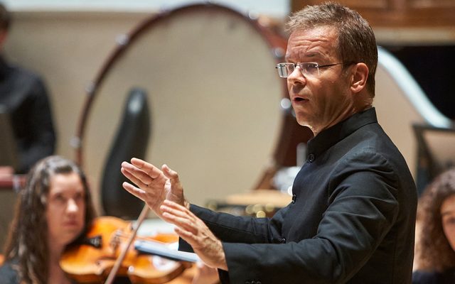 A man in a black shirt wearing glasses conducting an orchestra.