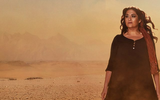 Image showing a woman, Aida, with long dark hair and a black dress is walking barefoot across a desert with black smoke behind her from what looks like bombing