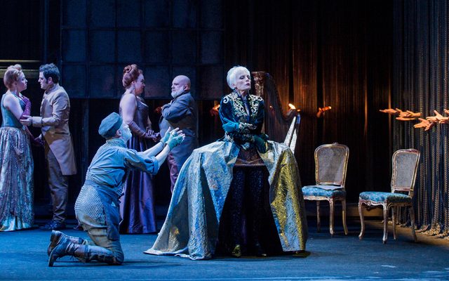 A woman in an elaborate gown stands looking away from a man kneeling. On the right some hands are reaching through some chains.