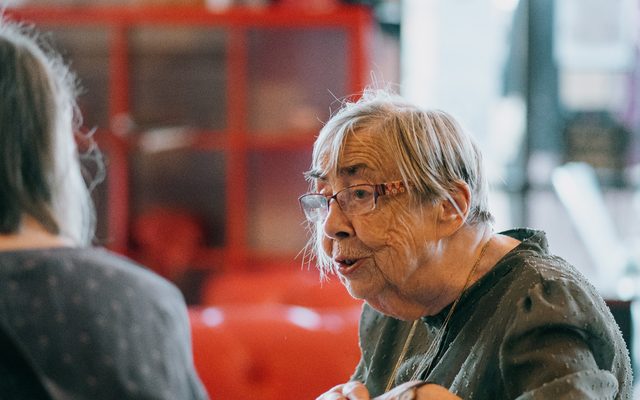 An older woman wearing glasses is talking to another woman.