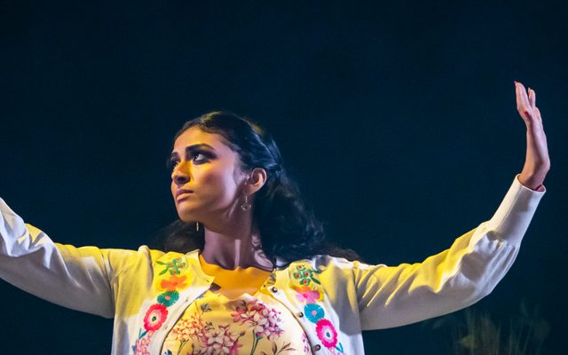 A woman wearing a white jacket with flowers on with her arms outstretched.