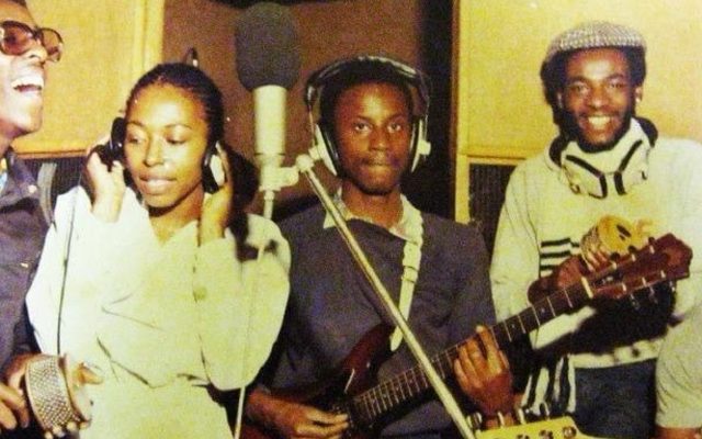 A reggae band and female singer gather around a microphone in a grainy vintage photo