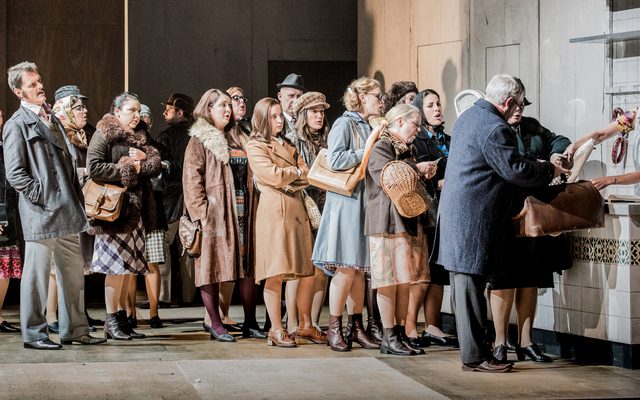 A wide shot of an opera. On the stage the chorus are queuing at a shop looking unhappy.