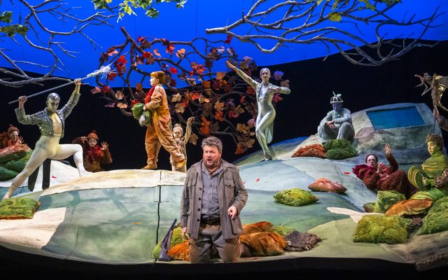The cast of The Cunning Little Vixen. They are sat on rolling hills with branches above them. The Forester is at the front of the stage and behind him there are people dressed as different animals.