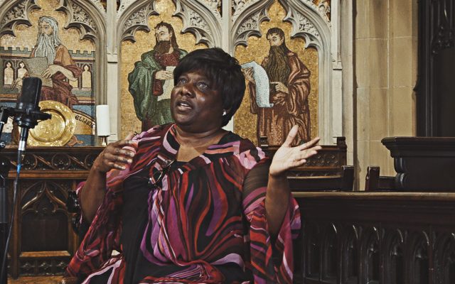 A woman in a brightly coloured dress gestures as she sings into a microphone, in front of an ornate gilt reredos