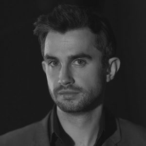 A man with dark hair wearing a suit with no tie.