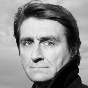 A man with short dark hair wearing a jacket with his collar raised.