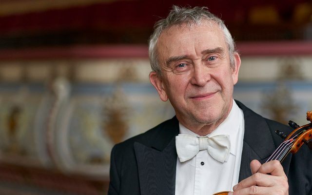A man in a bow tie and tails holds a violin, with an ornate gilt, red and blue stucco interior behind him