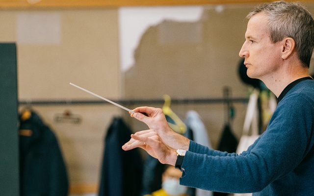 A man wearing a blue jumper conducting musicians.