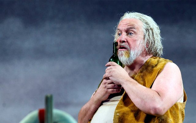 A man with grey hair and beard holding a wine bottle to his face looking forlorn.