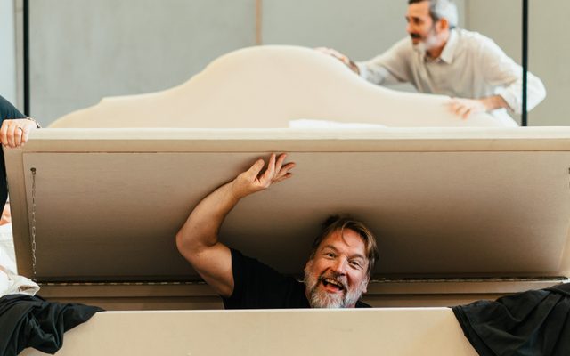 A white man with a beard and wearing a black top lifts up the lid of a laundry box where he's hiding at the end of the bed