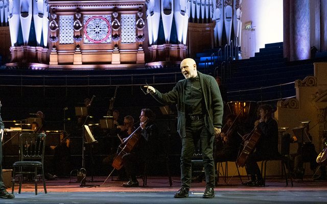 Singers in black stand on stage in front of an orchestra, and organ lit up.
