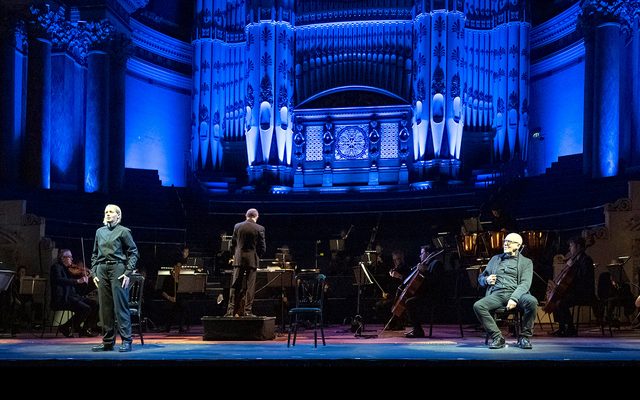 Singers in black stand on stage in front of an orchestra, and organ lit in blue.