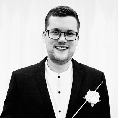 A man wearing glasses and smiling. He is holding a conductor's baton.
