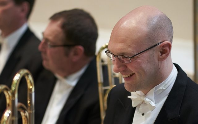 A smiling man in white tie and tailcoat with a trombone, surrounded by other brass players