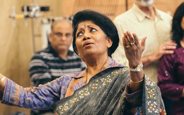 A woman in a traditional Indian sari raises her hands palms upwards as she sings