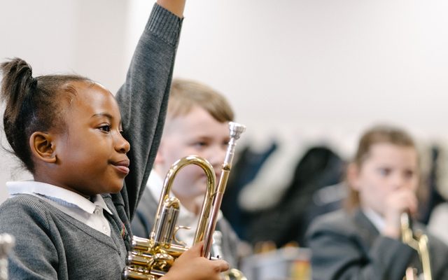 A young girl holds a trumpet in one hand with her other hand raised.