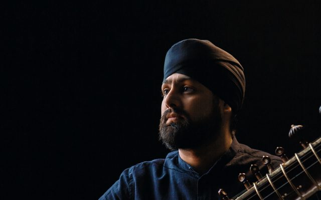 A man in a black shirt and turban, holding a sitar.