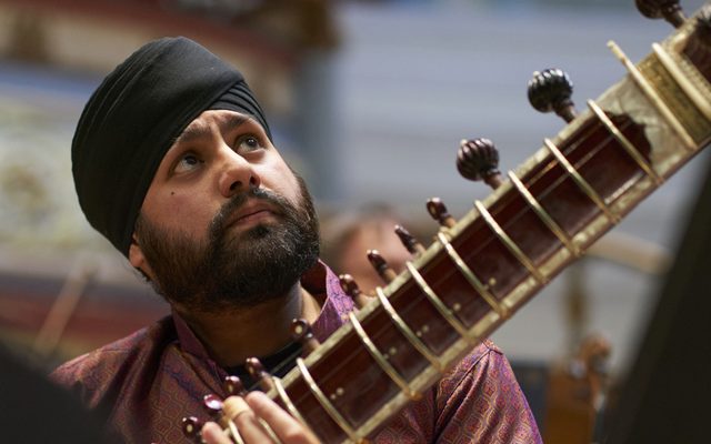 Sitting on a stage, a sitarist in a turban and patterned tunic looks up to his left