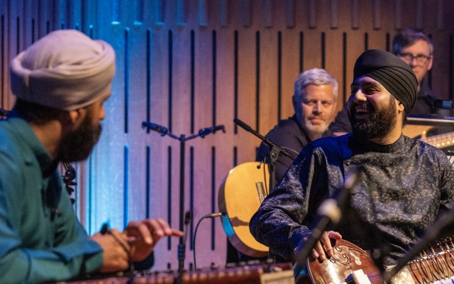 A group of musicians are on stage, playing instruments including a sitar and a santoor.