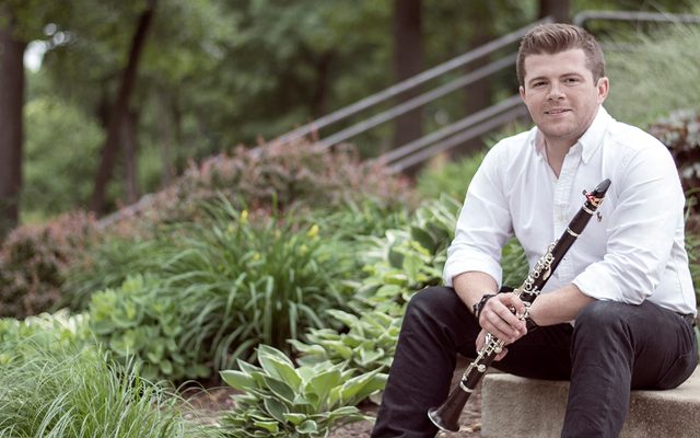 A man in a white shirt sat on some steps out doors holding a clarinet.