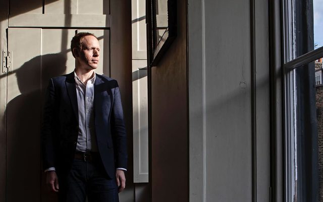 A man with short hair is stood looking at a window. he is wearing a suit without a tie.