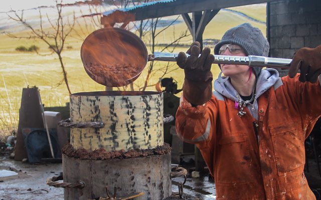 A woman in an orange boiler suit outdoors pours flaming molten metal into a mould