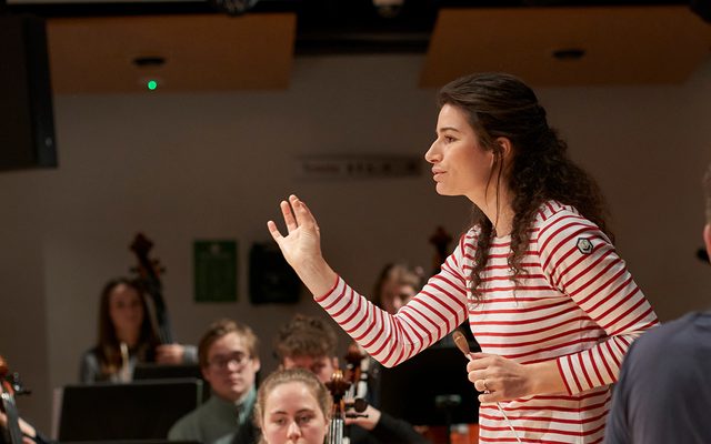 A young woman in a red and white striped jersey conducts an orchestra