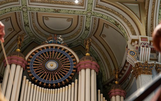An ornate pipe organ and coffered ceiling with a conductor's hand and baton and a cellist in the foreground on either side