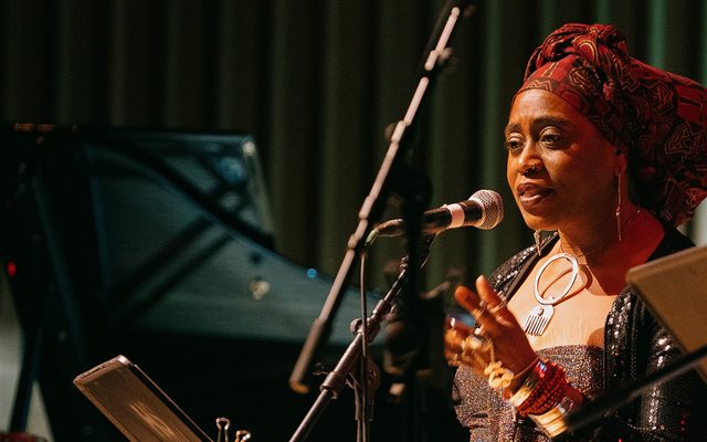A woman in headscarf and large silver necklace reads into a microphone