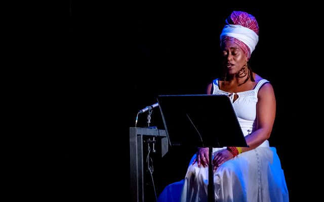 Poet Khadijah Ibrahiim, wearing a white dress and headscarf, performs seated on a darkened stage with a microphone and music stand