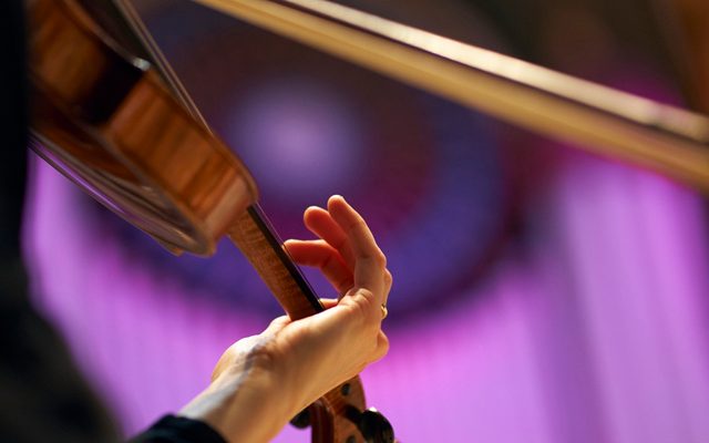 A musician's hand on the neck of a violin, the other hand holding a bow, with organ pipes lit in dramatic purple in the background