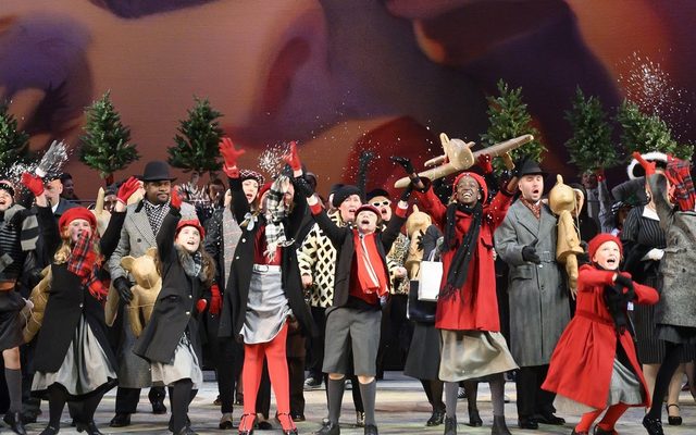 A crowd of children dressed in hats and scarves wave their Christmas presents in the air.