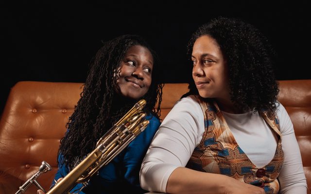 Two women sat on a sofa looking at each other. One is holding a trombone.