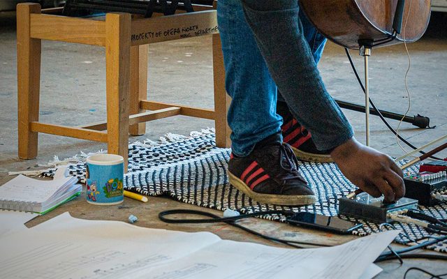 A cellist leans down to adjust an effects pedal on the floor, next to an outsized musical score