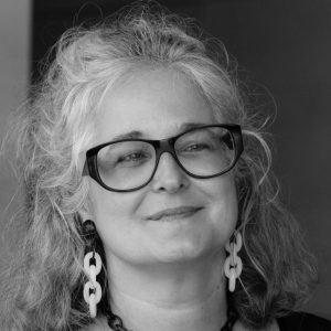A woman with long grey hair, thick rimmed glasses and dangling earrings.