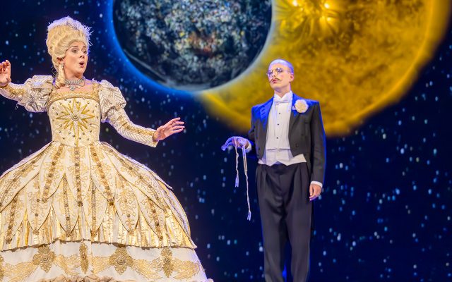 A woman in an ornate gold gown stood beside a man in a black suit. Behind them is a projection of the sun and earth.