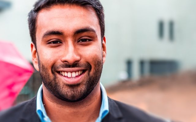 A man is looking at the camera smiling. He is wearing a light blue shirt and has a light beard.