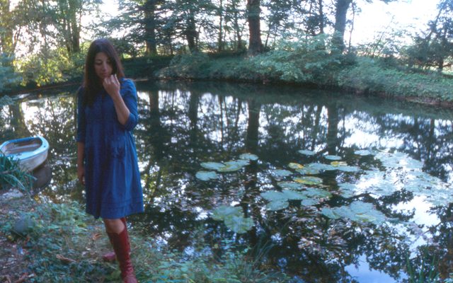 A woman wearing a blue dress and red boots next to a pond.