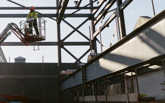 A workman on a hydraulic boom lift surrounded by an open steel frame, on a sunny day