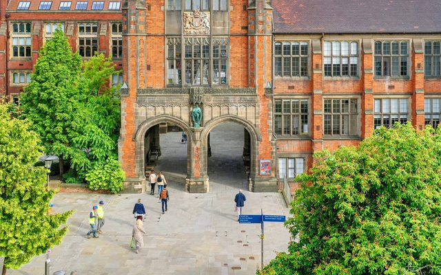 An aerial view of a courtyard dotted with trees in leaf, looking towards a redbrick building with two gothic arches at the bottom