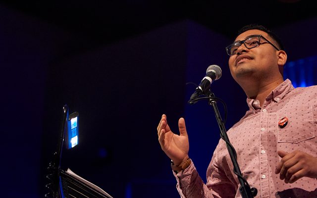 A man with glasses in a pink shirt is seen from below, gesturing with his hands as he sings into a microphone